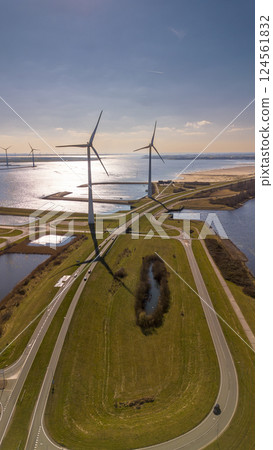 A scenic aerial view of a coastal wind farm with multiple wind turbines standing over the water. Curved roads lead through the landscape, with cars visible. The sun reflects off the water. A scenic aerial view of a coastal wind farm with multiple wind turbines standing over the water. Curved roads lead through the landscape, with cars visible. The sun reflects off the water. 124561832