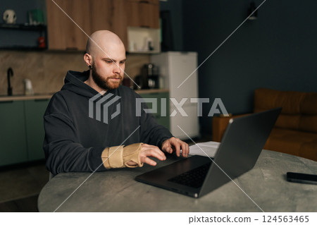 Concentrated freelancer male with wrist pain wearing orthopedic brace remote working on laptop computer from home office. Concept challenges of remote work and importance of ergonomics. 124563465