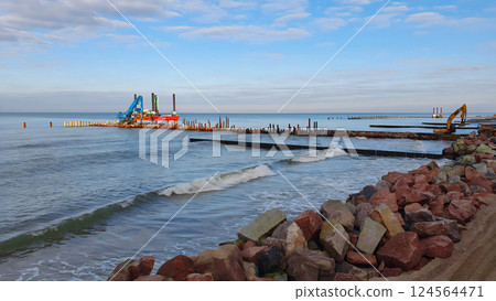 Construction machinery carries out shore protection works on stormy Baltic sea coast by laying large boulders and driving piles at sunny winter day. With no people seascape. Construction machinery carries out shore protection works on stormy Baltic sea coast by laying large boulders and driving piles at sunny winter day. With no people seascape. 124564471
