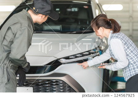 A male car mechanic and a female front desk clerk in office clothes having a conversation in front of a car 124567925
