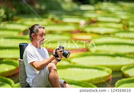 Asian Man is holding a camera and taking a photo at outdoor field with the Lily Lotus Leaf pond background. Asian Man is holding a camera and taking a photo at outdoor field with the Lily Lotus Leaf pond background. 124568610