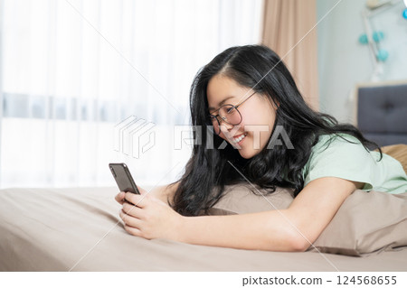 Beautiful glasses Asian woman lies on her stomach and plays mobile on the bed in her bedroom with a pastel green - brown color theme. Beautiful glasses Asian woman lies on her stomach and plays mobile on the bed in her bedroom with a pastel green - brown color theme. 124568655