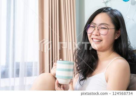 Asian Glasses Woman holds a cup of coffee and relax on the bed in the morning at her bedroom. Asian Glasses Woman holds a cup of coffee and relax on the bed in the morning at her bedroom. 124568734