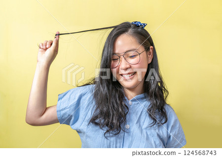 A young glasses and blue shirt asian girl touch her hair smoothly with a big smile on her face in front of yellow background. A young glasses and blue shirt asian girl touch her hair smoothly with a big smile on her face in front of yellow background. 124568743