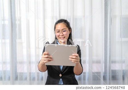 Smiling Asian Woman holds and watches gray tablet on her hand in front of curtain in private room in Work from Home period. Smiling Asian Woman holds and watches gray tablet on her hand in front of curtain in private room in Work from Home period. 124568748