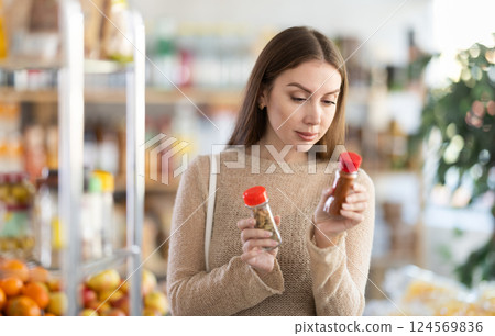 Positive girl standing in spice section of supermarket and choosing jar of spices or seasonings Positive girl standing in spice section of supermarket and choosing jar of spices or seasonings 124569836