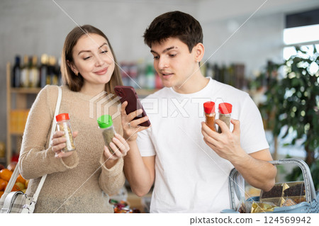 Beautiful young wife tells her husband what delicious spices and seasonings to buy, scans QR code from label in supermarket 124569942