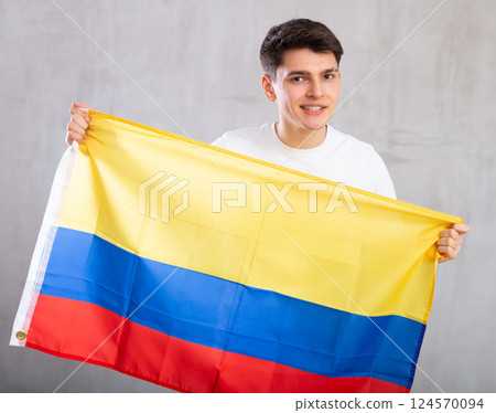 Man in holds unfurled flag of Colombia in hands raised above head against gray wall 124570094