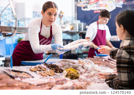 Female seller in apron standing near counter offering fresh fish striped red mullet to buyer 124570140
