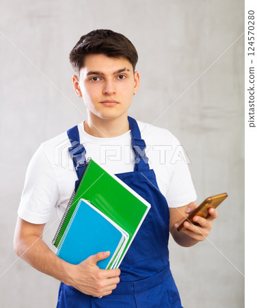 Young male builder in protective overalls with notebooks in his hands writes a message on smartphone screen Young male builder in protective overalls with notebooks in his hands writes a message on smartphone screen 124570280