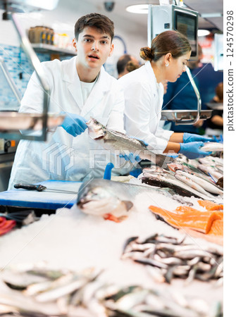 Male seller in apron standing near counter offering fresh fish hake to buyer 124570298