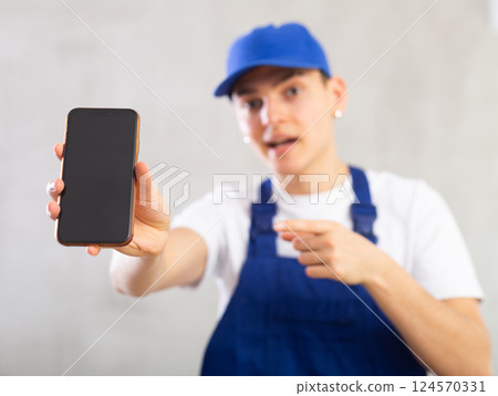 Unfocused man in jumpsuit and cap shows close up empty phone screen and points at device with finger 124570331