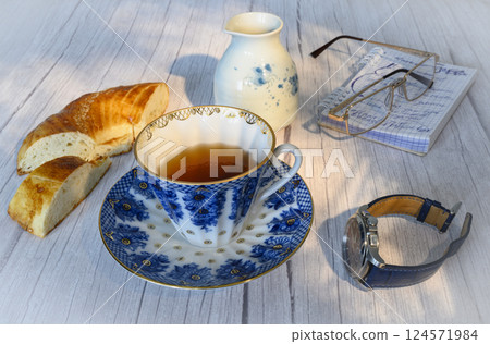 A cup of tea, a wristwatch and bread on the background of a jug, glasses and a notebook 124571984