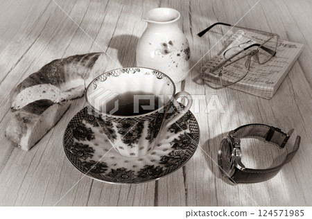 A cup of tea, a wristwatch and bread on the background of a jug, glasses and a notebook, in black and white A cup of tea, a wristwatch and bread on the background of a jug, glasses and a notebook, in black and white 124571985