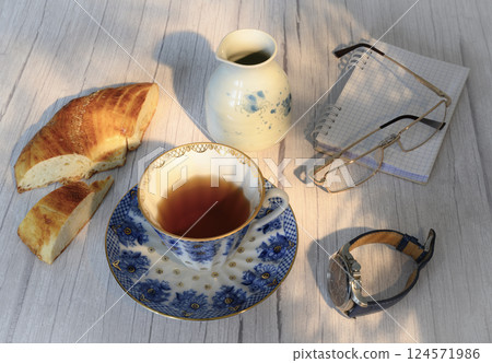 A cup of tea, a wristwatch and bread on the background of a jug, glasses and a notebook 124571986