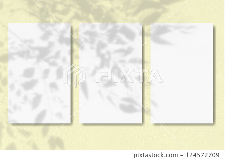 Natural light casts shadow from the leaves of a tree branch on 3 vertical sheets of textured white paper on yellow table background. Mockup 124572709