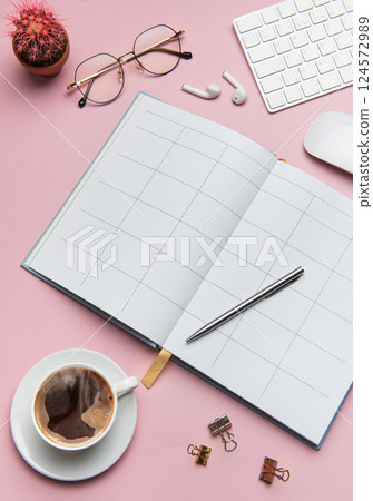 Open notebook, coffee, cactus, glasses, earphones, pen, keyboard and mouse lying on pink table 124572989