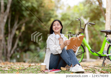 A woman looking happily at her notebook as she figure something out while sitting on park sidewalk 124574049