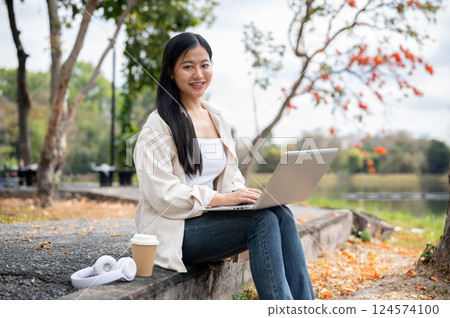 A woman looking happily at her notebook as she figure something out while sitting on park sidewalk 124574100