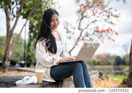 A woman looking happily at her notebook as she figure something out while sitting on park sidewalk 124574101