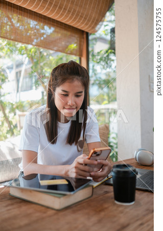 A young woman in a casual setting, using her smartphone at a cafe table with books and coffee, surrounded by a sunlit, modern interior. A young woman in a casual setting, using her smartphone at a cafe table with books and coffee, surrounded by a sunlit, modern interior. 124575755