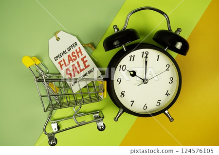 Flash Sale text on price tag with trolley shopping cart and alarm clcok top view on green geometric background 124576105