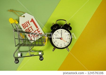 Buy 1 Get 1 free text on price tag with trolley shopping cart and alarm clcok top view on green geometric background Buy 1 Get 1 free text on price tag with trolley shopping cart and alarm clcok top view on green geometric background 124576106