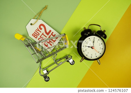Buy 2 Get 2 Free text on price tag with trolley shopping cart and alarm clcok top view on green geometric background 124576107