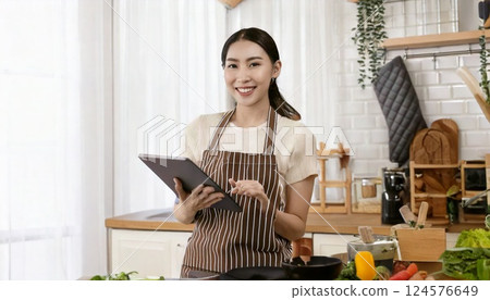 A smiling Japanese woman cooking while holding a tablet in one hand 124576649