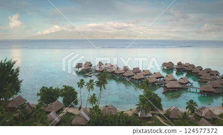 Breathtaking aerial view overwater bungalows in crystal clear turquoise water of French Polynesia, green vegetation and a distant island palm trees. Exotic travel vacation getaway, romantic honeymoon 124577372