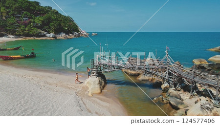 Aerial drone footage capturing a picturesque tropical beach with tourists strolling along the shore, a rickety wooden pier over turquoise waters, and lush greenery on the hillside 124577406