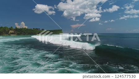 Stunning aerial view capturing turquoise ocean waves breaking on Dreamland Beach in Bali, framed by lush green cliffs and white clouds, creating a tropical paradise 124577530
