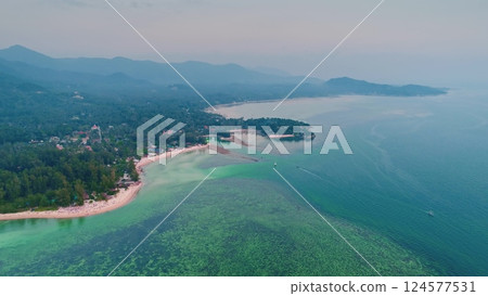 Aerial view of a tropical island coastline showing crystal clear turquoise water, lush green vegetation, boats sailing, and mountains in the distance 124577531