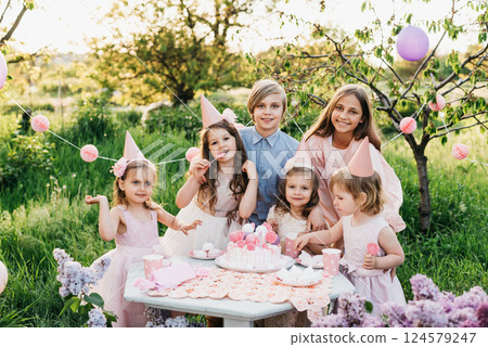Summer outdoor kids birthday party. Group of happy Children celebrating birthday in park. Children blow candles on birthday cake. Kids party pink pastel decoration and food. Presents and sweets. Summer outdoor kids birthday party. Group of happy Children celebrating birthday in park. Children blow candles on birthday cake. Kids party pink pastel decoration and food. Presents and sweets. 124579247