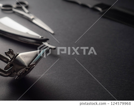 A variety of barber tools are displayed on a dark surface, including scissors, a comb, and a hair clipper. The arrangement showcases their shine and craftsmanship. A variety of barber tools are displayed on a dark surface, including scissors, a comb, and a hair clipper. The arrangement showcases their shine and craftsmanship. 124579968