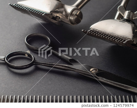 A collection of barber tools is displayed, featuring shiny clippers, sleek scissors, and a black comb resting on a dark surface. This setup emphasizes precision in grooming. 124579974