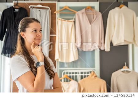 Young woman stylist in front of wardrobe filled with diverse clothing, analyzing outfit combinations. Creating fashionable looks requires attention to detail, color coordination, keen sense of style 124580134