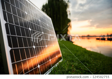 Solar panel illuminated by setting sun stands on grassy lakeside, with picturesque sunset and trees in background. Fusion of renewable energy technology with serene natural landscape. Solar panel illuminated by setting sun stands on grassy lakeside, with picturesque sunset and trees in background. Fusion of renewable energy technology with serene natural landscape. 124582755