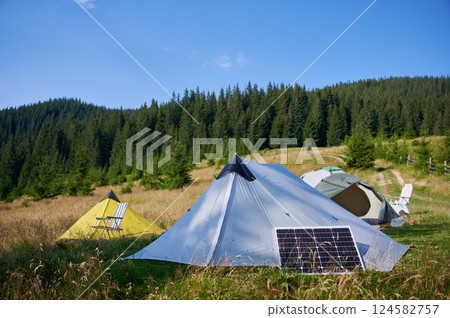 Three tents, including yellow one with striped chair, set up in grassy field with solar panel. Backdrop features lush forest and rolling hills under clear blue sky, creating tranquil camping scene. 124582757
