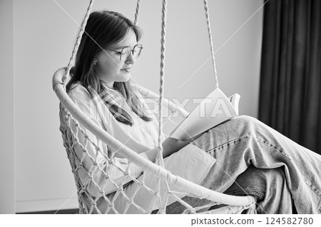 Beautiful girl relaxing and reading book on indoor swing. Pretty female in glasses excited about story she reading. Cozy afternoon with book in hanging chair in home. Black and white image. 124582780