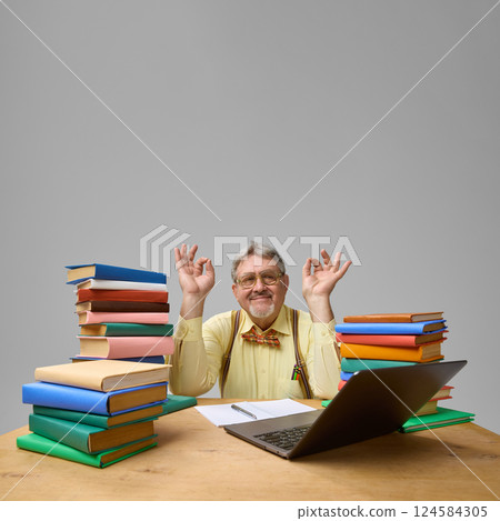 Professor meditating at desk with eyes closed, smiling, books and laptop around against grey studio background. 124584305