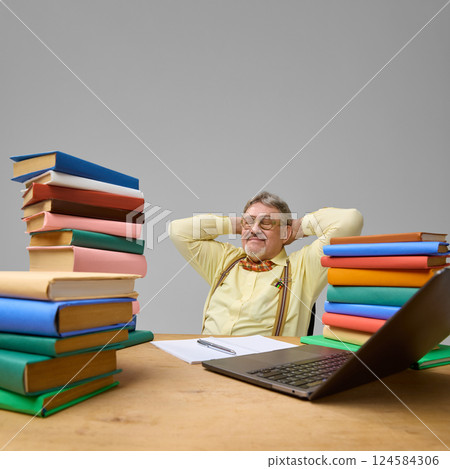 Senior man leaning back with satisfaction, hands behind head, surrounded by books against grey studio background. Senior man leaning back with satisfaction, hands behind head, surrounded by books against grey studio background. 124584306