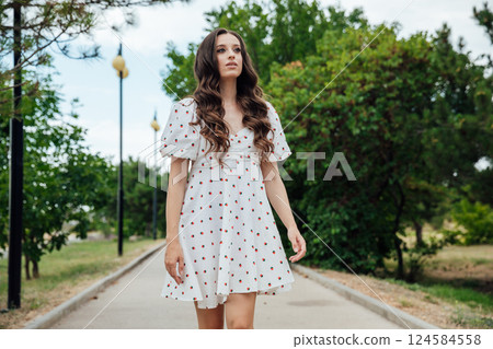 slim fashionable brunette woman in summer white dress walking along the alley in the park slim fashionable brunette woman in summer white dress walking along the alley in the park 124584558