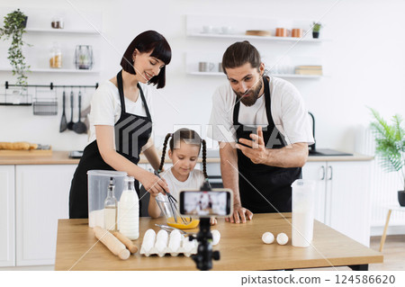 Caucasian family of bloggers filming cooking tutorial in kitchen. Parents teaching daughter baking. Whisking, kneading ingredients for dough in inviting atmosphere. 124586620