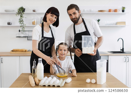 Caucasian adult man and woman with young girl cooking together in home kitchen, smiling and enjoying making food as a family while using eggs, milk, and flour. Caucasian adult man and woman with young girl cooking together in home kitchen, smiling and enjoying making food as a family while using eggs, milk, and flour. 124586630