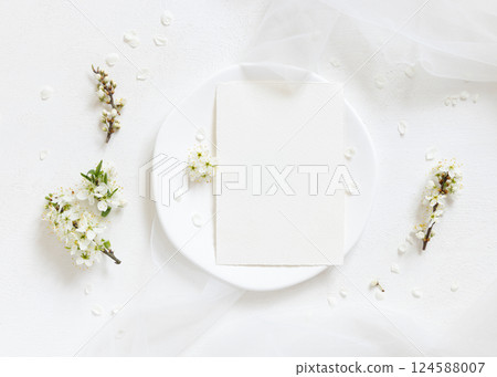 Blank card on plate near tulle and wild plums white flowers top view, romantic wedding mockup 124588007