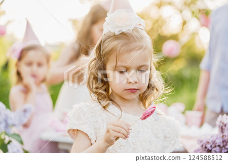 Summer outdoor kids birthday party. Group of happy Children celebrating birthday in park. Children blow candles on birthday cake. Kids party lilac pink pastel decoration and food. Presents and sweets. Summer outdoor kids birthday party. Group of happy Children celebrating birthday in park. Children blow candles on birthday cake. Kids party lilac pink pastel decoration and food. Presents and sweets. 124588512