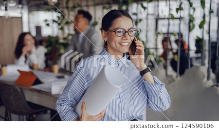 A Professional Woman in an Office Environment Engaging on the Phone with Detailed Blueprints 124590106