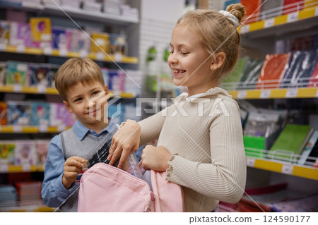 Little children putting new school supplies into backpack standing in store 124590177