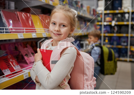 Portrait of smiling girl child carrying red copybook in hands posing in shop 124590209